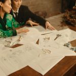 Dois designers trabalham em um projeto de moda cercados por esboços e ferramentas em um estúdio.